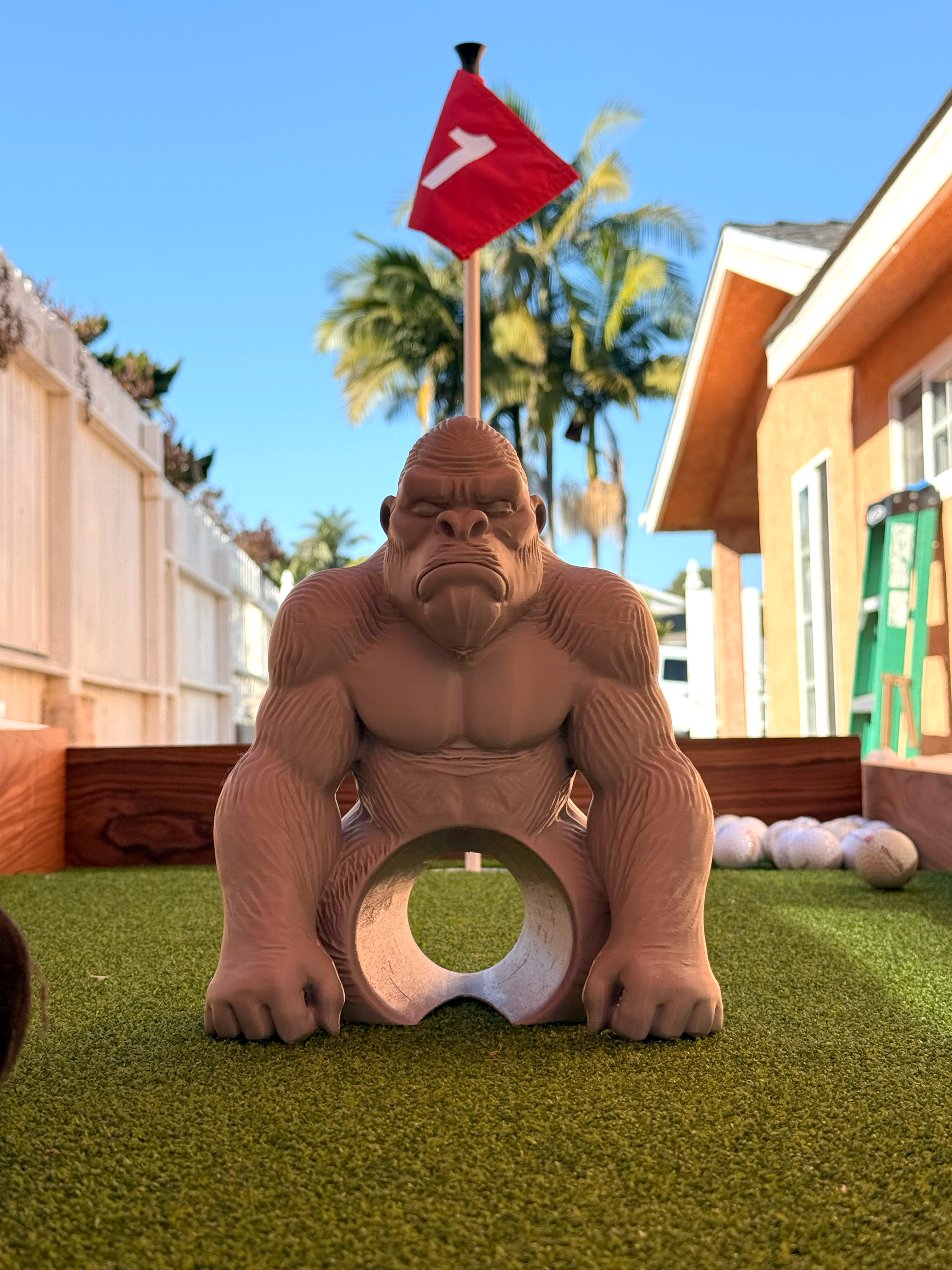 A gorilla-shaped miniature golf hole target on green artificial turf, with a red flag featuring a white number 7 in the background of an outdoor backyard setting.